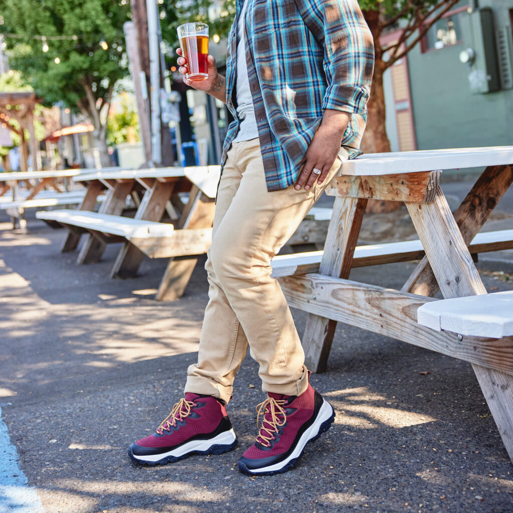 Man drinking a beer, leaning on a picnic table wearing khaki pants, a blue flannel and red hiking shoes